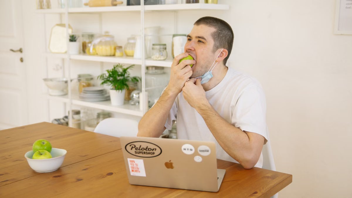 A man enjoys a green apple while sitting at a wooden table with a laptop during remote work.