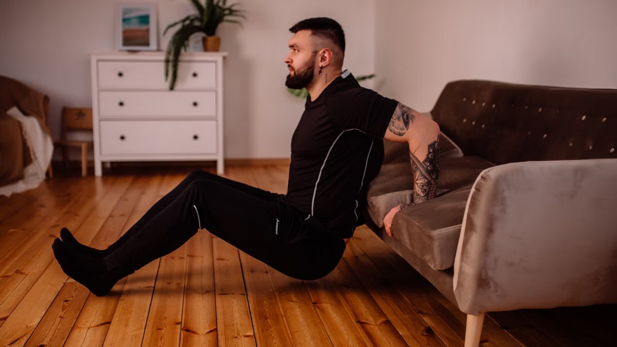 A bearded man in black fitness attire exercising at home, showcasing arm tattoos and a healthy lifestyle.